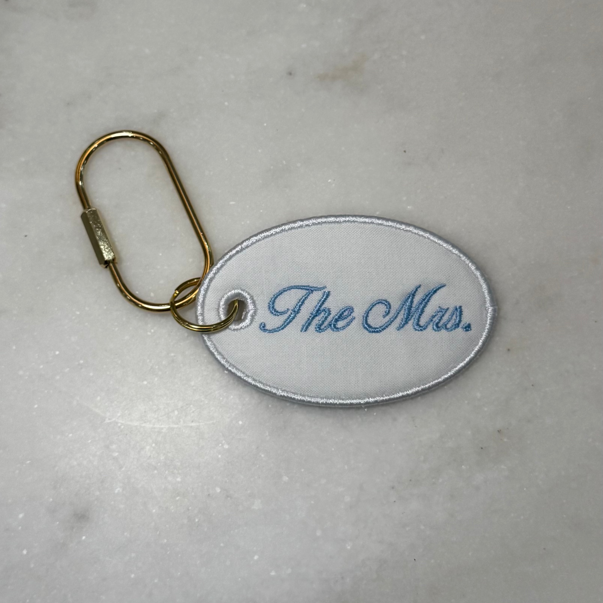 White oval keychain with 'The Mrs.' embroidery on a marble surface