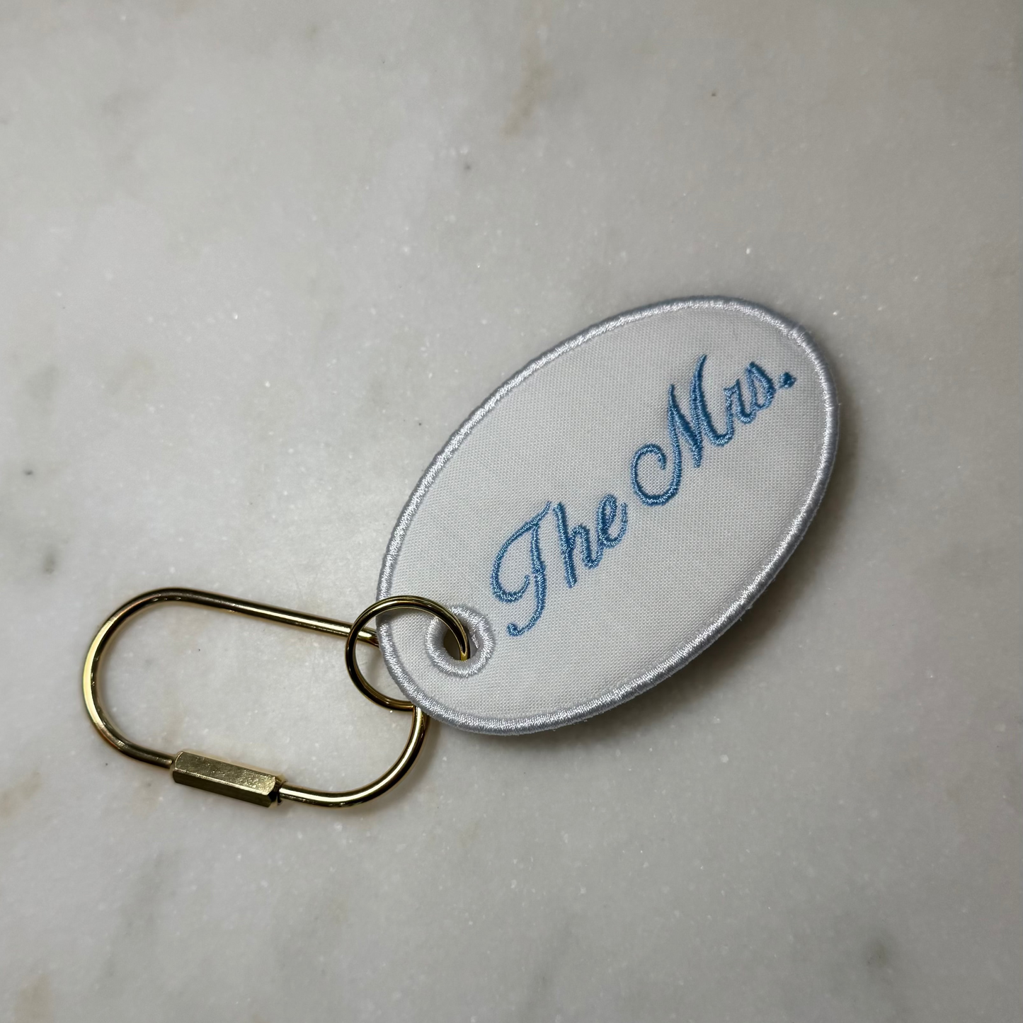 Oval key tag with 'The Mrs.' embroidery on a beige background